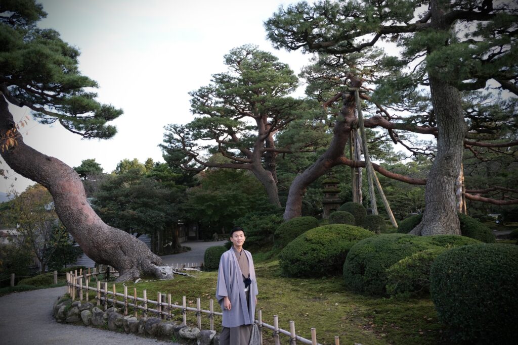 Steven in traditionele kimono staat in een prachtige Japanse tuin. De omgeving is rustgevend, met zorgvuldig gesnoeide pijnbomen en mosheuvels die de tijdloze schoonheid van de natuur laten zien.